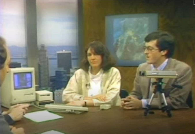 A demonstration of the Magic video digitizer. A woman and a man are sitting at a desk. On the desk, there is a small security camera mounted on a tripod hooked up to an original Apple Macintosh computer.