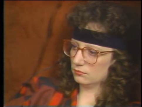 A woman with dark curly hair and glasses seated in a chair. She is wearing a large black headband and has a bored expression on her face.