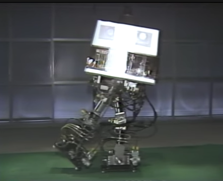 A robot with a cubic head and four motorized limbs walking across a stage.